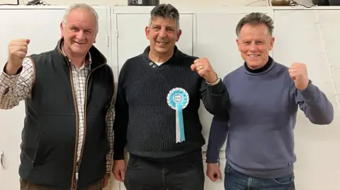 BBC Heinz Glanville wearing a dark jumper with a Reform rosette on, standing between Paul Ashton in a checked shirt and black bodywarmer, and Roger Tarrant in a grey jumper and jeans. They are all raising their fists in celebration and stand in front of some wide doored cupboards. 