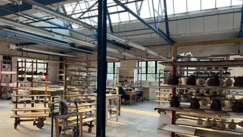 Supplied An empty pottery studio with workbenches, drying racks, clay pots, trolleys and shelves. There is strip lighting turned off and sun beams through the far left windows.