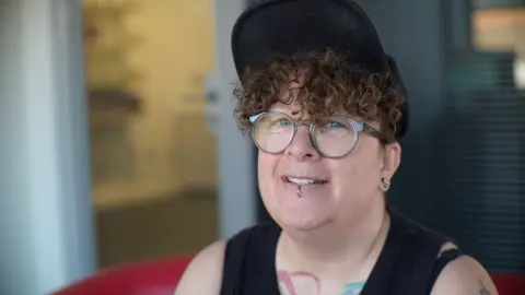 Jamie Niblock/BBC Emma Hastings looks at the camera. She has brown curly hair, wears a cap and glasses and has a lip piercing.