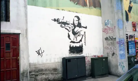 Getty Stencil street art on a white wall depicting a woman resembling the Mona Lisa holding a rocket launcher, surrounded by graffiti tags and set above two utility boxes on an urban street.