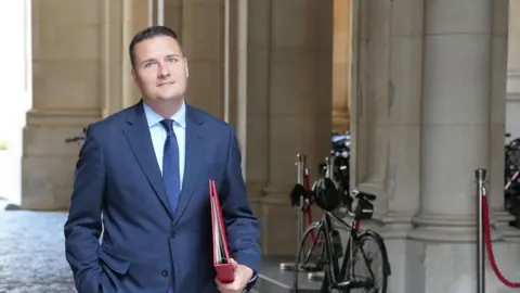 Wes Streeting walking down the street. He is in a navy suit and carrying a red folder.