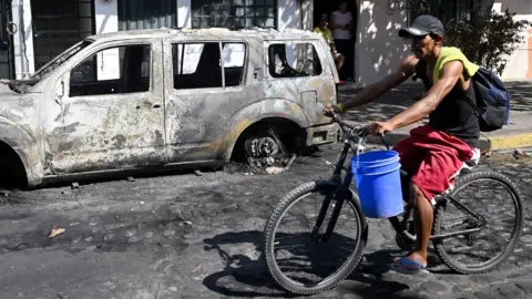 A man rides a bicycle past a burned vehicle in Puerto Vallarta, Jalisco State, Mexico, on February 24, 2026.