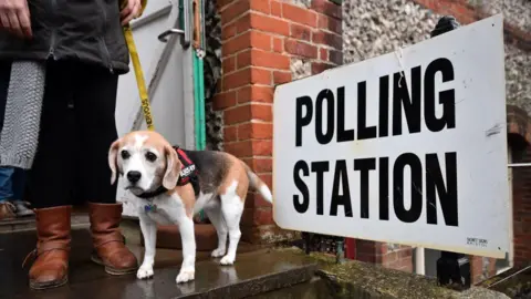 A voter wearing brown boots and a long grey cardigan leaves a polling station in Brighton with a small brown dog on a yellow leash. 