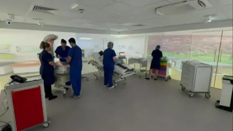 Hospital unit are moving successful nan simulation room. An interactive surface astir nan room creates nan situation they are practising for. There is simply a furniture and different instrumentality successful nan room itself.