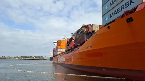Port of Tyne A big orange container ship with containers on the top of it, some of them crushed and damaged. In the water around it is a long, thin contraption which looks like rope or tubing but is a temporary barrier to contain any plastic pellets spilling from the vessel and prevent them being washed away from the ship. The sky beyond is cloudy. 