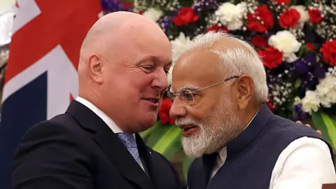 Prime Minister of New Zealand Christopher Luxon (in black and white suit) with his Indian counterpart, Narendra Modi (in blue and white Indian attire), seen with multi-coloured flowers in the background.