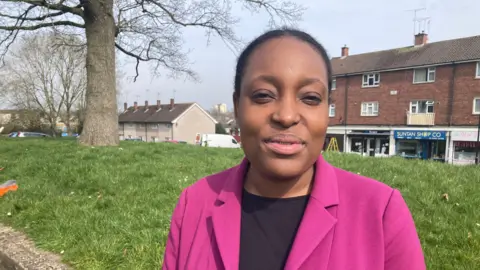 A woman wearing a pink smart jacket and a black top underneath. She is smiling at the camera. We can see a green grassy bank behind her as well as a number of shops and houses. It is a bright but overcast day.