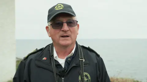 Jerry Betteridge stands in front of the coastline. There is a grey November sea behind him and the edge of a white wall of the lookout post to his side. He has a camera strap around his neck.