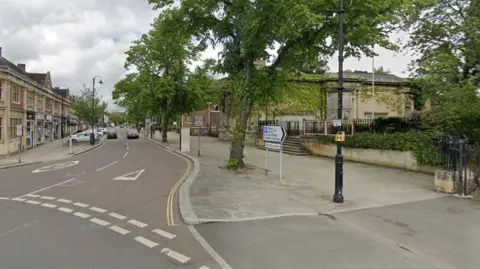 Google Town centre road junction showing painted Give Way symbol and 20MPH sign on the road. There is also a roadsign next to a large tree directing motorists to a car park. There is a stone wall topped with foliage alongside steps to the right, and two-storey shops to the left. 