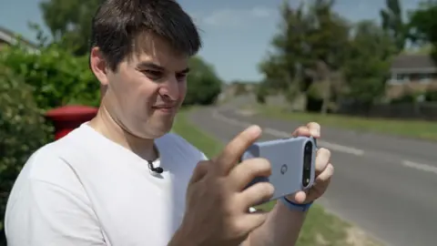 A man in a white t-shirt stands by the side of a road and holds up his phone, looking at the screen. In the background there is a residential road curving in to the distance, lined with trees and grass.
