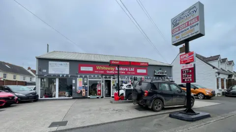 A view of the outside on the petrol station showing its red sign. Cars are for sale to the left and cars are parked at the two pumps. 