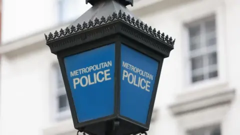 A Metropolitan police lamp post which is blue inside with the Metropolitan Police words in white writing. The post itself is black metal. There's a blurry white building in the background 