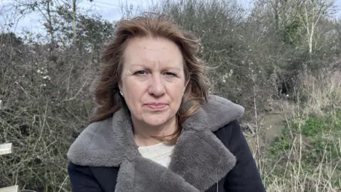 BBC/ Kate Bradbrook A woman with brown hair, wearing a cream jumper and black coat with grey furry collar stands in front of bramble and a brook