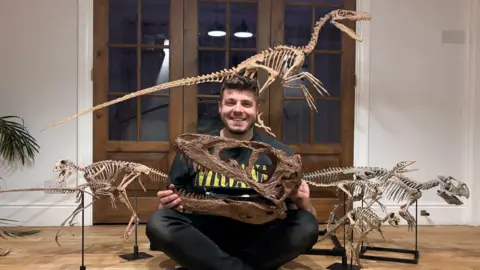 Alex Pritchard Mr Pritchard sits crossed leg on the floor, smiling at the camera, while holding the replica of a dinosaur skull. Small full replicas of other dinosaurs are placed either side of him and one behind him.