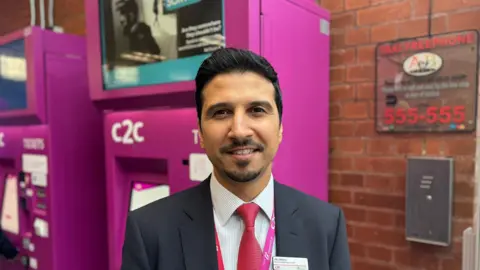 George King/BBC A man with short black hair and some facial hair wearing a suit standing in front of a pink ticket machine