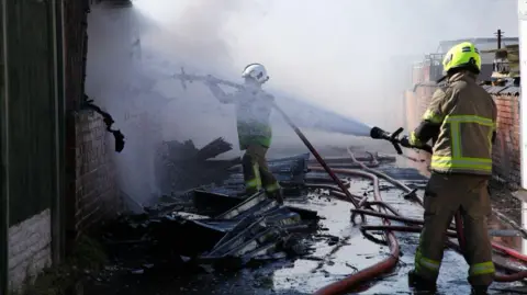 South Yorkshire Fire and Rescue Service Two firefighters working in a narrow alleyway between brick buildings that have been badly damaged by fire. The firefighters are wearing protective gear and helmets while using a hose to spray a powerful stream of water onto charred walls and smouldering debris. Burned materials, twisted metal, and pooled water cover the ground, with fire hoses running along the alley. Smoke and water mist fill the air.