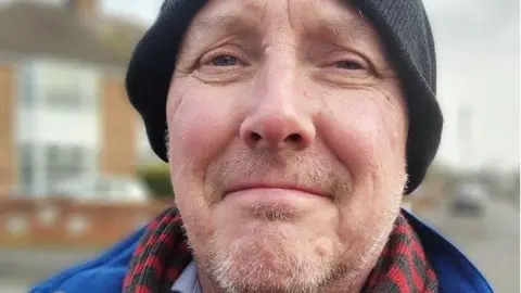 A man wearing a blue coat, red and grey scarf and black woollen hat looking at the camera