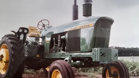 Dave Harrison A large metal green-bodied John Deere tractor with a yellow open air seat and yellow alloys.