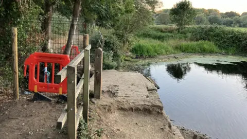 An image of a round pond, to the right of the image, with a meadow and trees beyond. To the left is a path leading off into the distance. Red plastic fence panels block the path