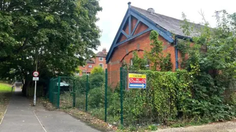 A church hall building, behind fencing which is covered with greenery. A cycle path runs past the near side of the fence.