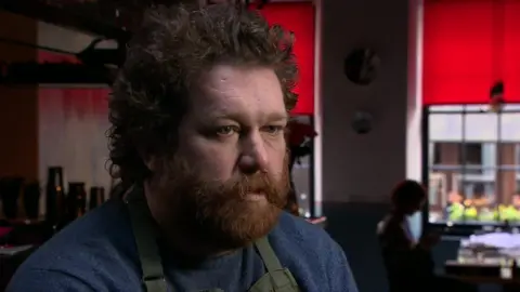 Alexei Zimin has a big bushy brown beard and long curly hair. He is looking towards the interviewer at an angle during an interview with the BBC in 2017 inside his Soho restaurant