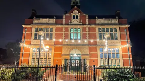 A night-time view of the Applecart Arts venue, located in the historic Passmore Edwards Building, a red-brick former library with large windows and decorative stone details, illuminated by string lights and street lamps.