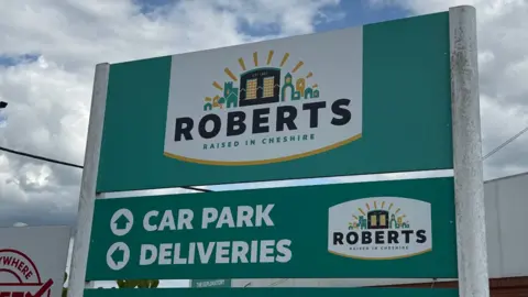 A turquoise-green sign with "Roberts: Raised in Cheshire" and car park and deliveries directions written on it