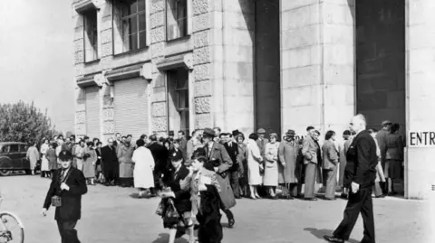 Droitwich Spa Heritage Centre A black and white photo of about 50 people queuing outside a stone building. It is a mix of men, women and children, all dressed smartly in long coats and hats.