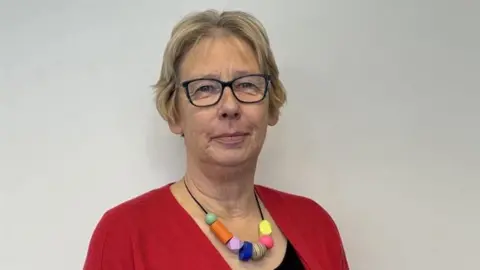 A woman with short blonde hair, glasses, a multi-coloured necklace, a red cardigan and black top, stands in front of a white wall looking at the camera.