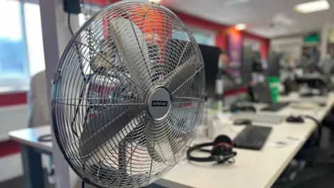 A metal, electrical fan in an office. A row of computers and keyboards are visible in the background.