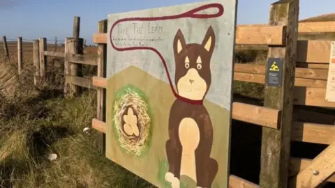 A sign nailed to a wooden fence warning to keep dogs on a lead, in the middle of a grass field. It shows a painting of a cartoon dog with a lead around its neck, next to a bird's nest with eggs in it.