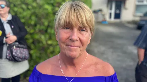 A woman is wearing a blue top with a silver chain. She has blonde hair. 