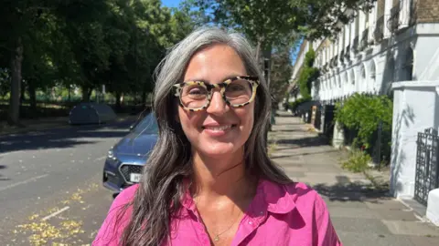Woman with tortoiseshell framed spectacles, grey and white long straight hair, and a bright pink shirt stood on a pavement in a residential area. She smiles at the camera.