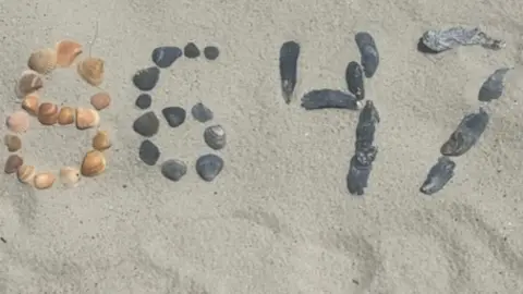 Close-up shot of sand with a bunch of shells and rocks spelling out 86 and 47