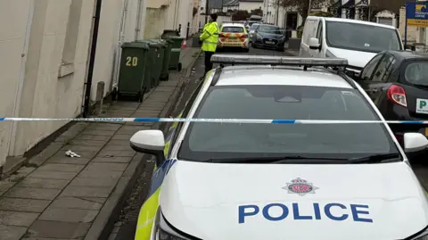 A police car parked close to the camera in the St Pauls area of Cheltenham. In front of it is a line of blue and white police tape stretched across the road. In the background a lone police officer in a high-vis top can be seen, along with some black wheelie bins