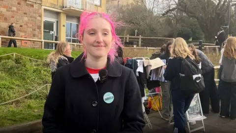 Annie McNamee is standing in front of a university building. There are people in the background standing around. Annie has pink hair and is wearing a black coat.