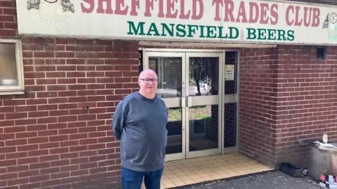David Spereall/BBC A bespectacled man stands outside a brick-built building, with a sign above the door saying 'Sheffield Trades Club, Mansfield Beers'. 