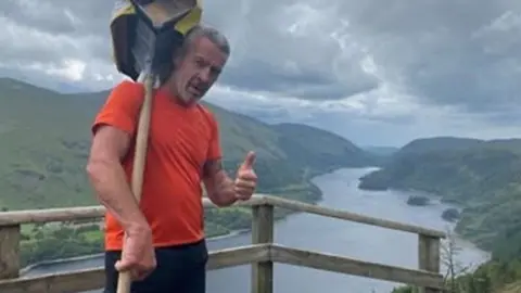 A man in an orange top standing on a viewpoint overlooking the lade district and a river. he is holding a long wooden stick with bricks attached to the top and has his thumb up