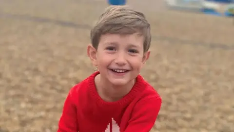 Young boy, Benedict, wearing a bright red jumper looks towards the camera outdoors, with a softly blurred background creating a natural portrait.