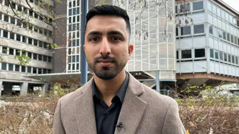 BBC Abdullah Tanoli stands outdoors in front of an office building in Croydon, wearing a brown jacket over a black shirt, with trees and modern buildings in the background.