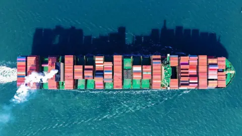 Getty Images This aerial picture shows a container ship carrying containers of various colours sailing out of Qingdao port, in China's eastern Shandong province. All around it are blue waters reflecting the shadows of the containers.     