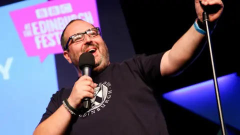 Justin Moorhouse on stage. He is holding a microphone and wearing a black t-shirt. He's wearing black glasses and smiling. 
