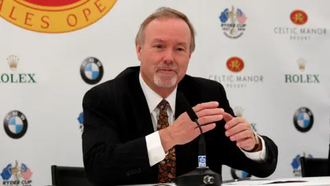 Getty Images Terry Matthews sitting at a desk in a press conference. He is wearing a black suit jacket with a white shirt and brown tie with leaves and plants on it. He has a grey goatee and short brown hair. Behind him is a white background with logos for BMW, Rolex, the Ryder Cup and Celtic Manor.