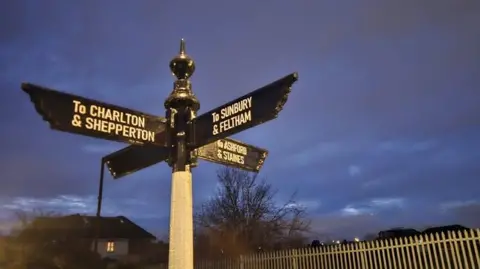 BBC Black finger signage pointing in four different directions. The signs are on a white post. The sky is dark grey behind the signs