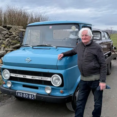 A man with white hair leans on a vintage blue Opel truck. It has a mulberry-coloured car on the back of it. 