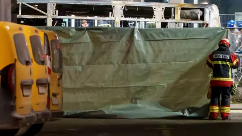 A firefighter standing to the right of the picture sets up a green barrier in front of the charred shell of the bus.
