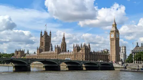Getty Images A wide view across the Thames shows the Palace of Westminster and the Elizabeth Tower, commonly known as Big Ben, against a blue sky with white clouds. Westminster Bridge stretches across the foreground with traffic and pedestrians crossing. The iconic clock face is clearly visible near the top of the tower.
