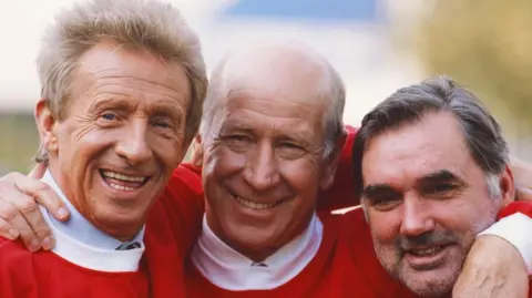 Getty Images Three men wearing red and white football strips stand with their arms around each other smiling at the camera. Denis Law, fair left, has light brown hair. Bobby Charlton has thinning grey hair and George Best has dark hair.