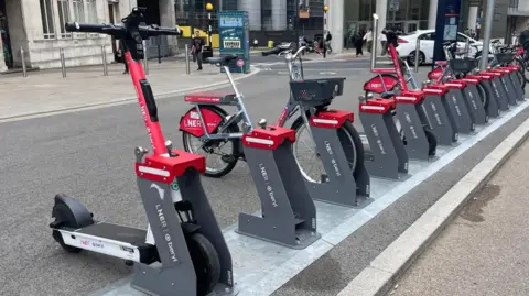 Red and white e-scooters and e-bikes at a docking bay in the city. They have LNER branding on the sides of them.