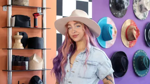 A woman with long hair which is dyed pink at the front, purple towards the back, looks towards the camera while wearing a cream, wide-brimmed hat. She wears a light blue short-sleeved denim top. Behind her are a variety of hats hanging on the wall or on shelves.